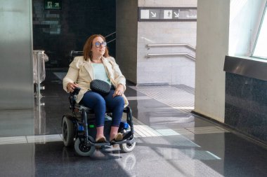 Elektrikli sandalyeyle ulaşılabilir bir metro istasyonunda gezen kendine güvenen iş kadını, şehir yaşamında bağımsız hareketliliği vurguluyor. Katılım kavramı, uyarlanmış toplu taşıma