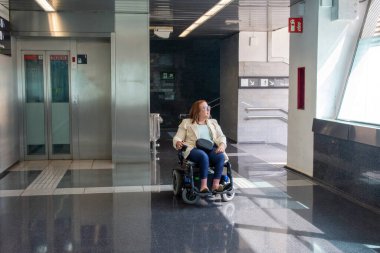 Açık bej takım elbiseli ve kot pantolonlu bir iş kadını modern bir metro istasyonunda elektrikli sandalyede oturuyor. Katılım kavramı, uyarlanmış toplu taşıma