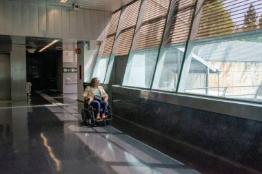 Üst düzey bir kadın parlak bir metro koridorunda bekliyor. Büyük pencerelerden içeri güneş ışığı altında modern bir elektrikli sandalye kullanıyor. Katılım kavramı, uyarlanmış toplu taşıma