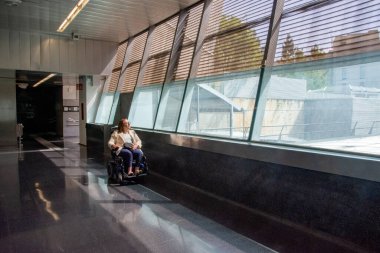 Tekerlekli sandalyedeki kadın, aydınlık ve modern bir metro istasyonunda geniş bir pencereden manzaranın keyfini çıkarıyor. Erişilebilirliği ve dahil edilmeyi ön plana çıkarıyor. Katılım kavramı, uyarlanmış toplu taşıma