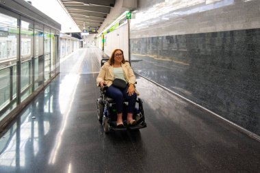Elektrikli sandalyesini kullanan mutlu iş kadını işe giderken modern bir metro istasyonunun içinde geniş bir koridor boyunca ilerliyor. Katılım kavramı, uyarlanmış toplu taşıma