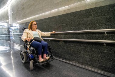 Tekerlekli sandalyede modern bir metro koridorunda gezinen gözlüklü bir iş kadını toplu taşımacılığa erişimi ve dahil edilmeyi vurguluyor. Katılım kavramı, uyarlanmış toplu taşıma