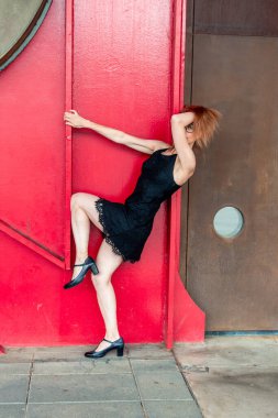Gösterişli siyah elbiseli, şık topuklu bir kızıl saçlı dansçı modern bir binanın çarpıcı kırmızı metal duvarına nazikçe poz veriyor ve bacaklarını esnetiyordu. Zarafet ve yaratıcılık saçıyordu.