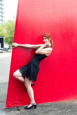 Siyah elbiseli kızıl saçlı dansçı şehir ortamında büyük kırmızı bir duvara yaslanmış canlı bir kontrast yaratıyor ve zarafet ve tarzını sergiliyor.