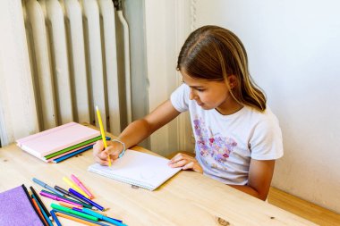 Evdeki ahşap masaya renkli kalemlerle çizen kız.
