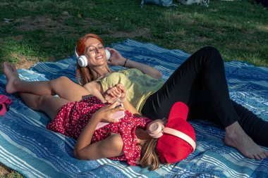 Anne ve kızı parkta bir battaniyenin üzerinde uzanmış, kulaklıkla müzik dinlerken, güneşli bir günün sıcaklığının tadını çıkarırken ve birlikte değerli anların tadını çıkarırken el ele tutuşuyorlar.