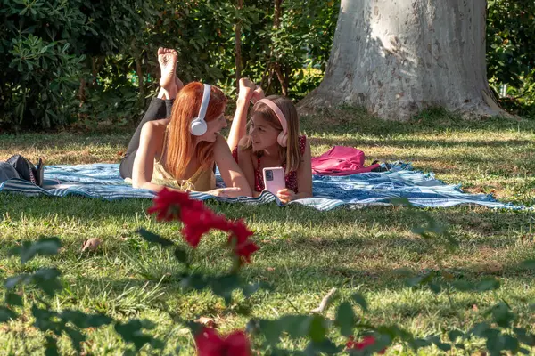 Mutlu anne ve kız birlikte güzel vakit geçiriyorlar, parkta bir battaniyeye uzanıp, kulaklık takıp, doğa ve güneş ışığıyla çevrili müzik dinliyorlar.