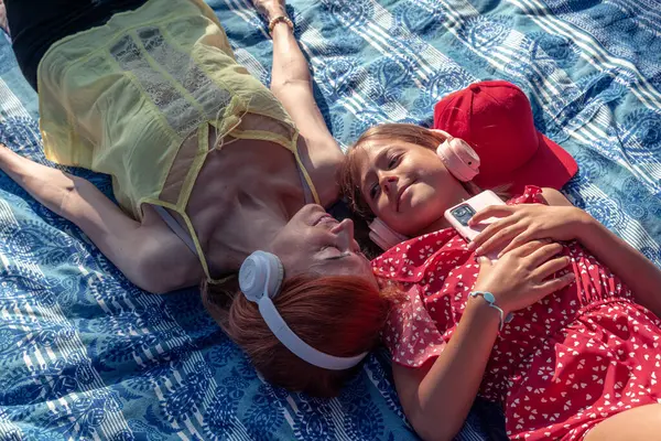 Kızıl saçlı anne ve kız kulaklık takıyor mavi battaniyeye uzanıyor aile pikniğinde müzik dinliyor, akıllı telefon tutuyor ve birbirlerine bakıyorlar.