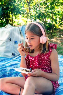 Parkta bir battaniyenin üzerinde oturan, kulaklık takan genç bir kız yaz pikniği sırasında yanında sırt çantasıyla müzik dinlerken atıştırmanın keyfini çıkarıyor.