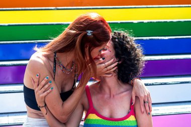 İki kadın, gökkuşağı renginde boyanmış merdivenlerde tutkulu bir öpüşme paylaşıyor. Gurur, sevgi ve eşitliği simgeliyor.