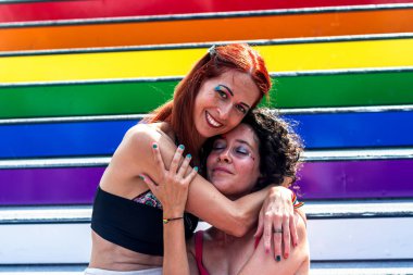 İki kadın birbirlerine renkli bir gökkuşağı bayrağıyla sarılıyorlar. Arkalarına da sevgi, şefkat ve lgbtq artı haklar ve toplum için destek yazıyorlar.