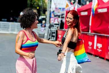 İki genç kadın el ele tutuşuyor ve gülümsüyor, bir geçit töreninde aşkı ve kapsayıcılığı kutluyorlar, gökkuşağı renkleri giyiyorlar ve mutluluklarını ifade ediyorlar.