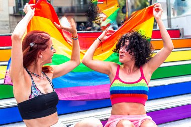 İki mutlu kadın gökkuşağı renkli tribünlerde oturuyor, ellerinde büyük bir gökkuşağı bayrağı sallıyorlar, eşcinsel gurur etkinliği sırasında gurur ve çeşitliliği kutluyorlar.