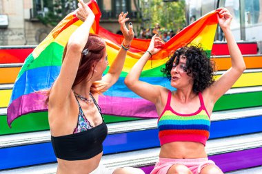 İki kadın gökkuşağı renkli tribünlerde oturmuş, ellerinde büyük bir gökkuşağı bayrağı, lgbtq 'ı kutluyorlar artı açık havada gurur ve çeşitliliği.