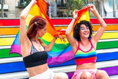 İki neşeli kadın gökkuşağı renkli tribünlerde oturuyor, ellerinde büyük bir gökkuşağı bayrağı sallıyor, lgbtq artı gurur ve çeşitliliği kutluyorlar.