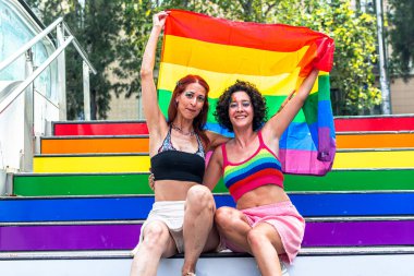 İki kadın gökkuşağı renkli merdivenlerde oturmuş, ellerinde büyük bir gökkuşağı bayrağı, gurur ve lgbtq artı hakları kutluyorlar, çeşitliliği, aşkı ve eşitliği sembolize ediyorlar.