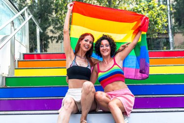 Igbtq artı gururu kutlarken ve çeşitlilik ve toplum ruhunu kucaklarken gökkuşağı renkli renkli merdivenlerde iki neşeli kadın ellerinde büyük bir gökkuşağı bayrağı tutuyorlar