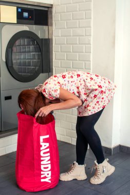 Genç bir kadın, çamaşırhanedeki büyük kırmızı bir çamaşır torbasını kontrol ediyor, çamaşırlarını yıkamaya hazırlanıyor ve günlük çamaşır işlerini hallediyor.