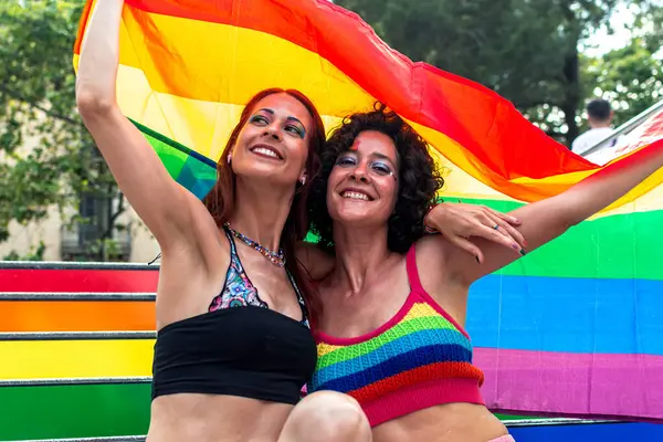 İki kadın büyük bir gökkuşağı bayrağını kucaklıyor ve tutuyor, Igbtq 'ı sembolize ediyor, artı gurur, çeşitlilik ve açık hava kutlamalarında ifade özgürlüğü.