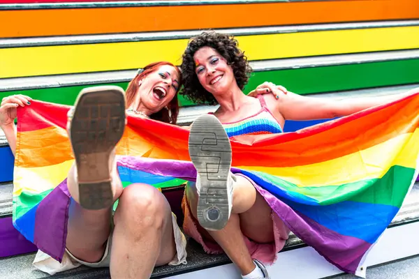 İki kadın ellerinde gökkuşağı bayrağı, renkli merdivenlerde oturmuş cinsel yönelimleriyle gurur ve sevinçlerini dile getiriyorlar.