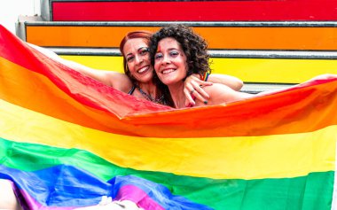 Gururla kucaklaşan iki neşeli kadın canlı, renkli adımların önünde büyük bir gökkuşağı bayrağı tutarken, gurur, çeşitlilik ve lgbtq artı haklar için mücadele ederken,