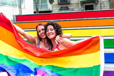 İki kadın gökkuşağı renkli merdivenlerin önünde büyük bir gökkuşağı bayrağını kucaklıyor ve taşıyorlar. Bu bayrak onların eşcinsel gurur etkinliğine katılımlarını ve lgbtq artı hak ve görünürlüğü kutlamalarını simgeliyor.