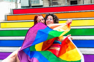 İki kadın gökkuşağı renkli merdivenlerde oturuyor, büyük bir gökkuşağı bayrağını kucaklıyor ve tutuyorlar, gurur verici bir olay sırasında lgbtq artı kimlikleriyle gurur ve neşe ifade ediyorlar.