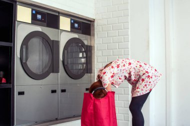 Çamaşırhanedeki çamaşır makinesine giysilerini koyup yükleyen genç bir kadın gerekli ev işlerini yapıyor.