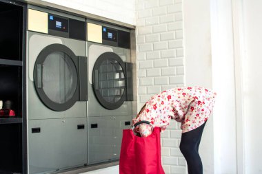 Genç bir kadın çamaşırhanedeki çamaşır makinesine giysilerini eğip yüklüyor. Çamaşırhanelerin kolaylığını ve verimliliğini vurguluyor.