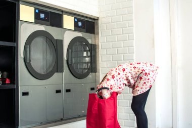 Genç bir kadın, modern bir çamaşırhanede giysilerini büyük bir kapasite kurutucusuna yüklerken eğiliyor. Duvarları parlak, beyaz fayanslı, temiz ve yeni bir çamaşır ortamı yaratıyor.