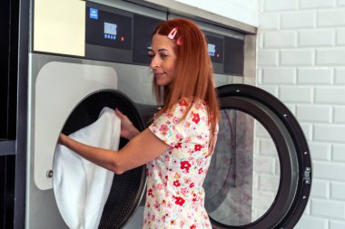 Kızıl saçlı kadın çamaşırhanedeki büyük bir çamaşır makinesinden beyaz havlu çıkarıyor ev işlerini yapıyor ve elbiselerini temiz tutuyor.