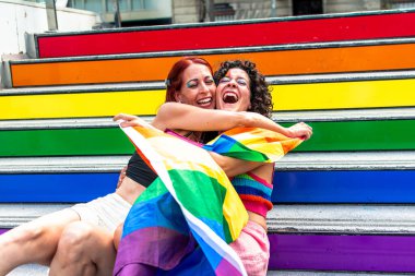 Gökkuşağı renkli renkli merdivenlerde sarılıp gülen iki kadın gökkuşağı bayrağına sarmalanmış gururu kutluyor ve lgbtq artı hakları ve kabulü savunuyor.