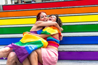 Gökkuşağı renkli canlı merdivenlerde oturan iki kadın, gökkuşağı bayrağına sarılmış, sıcak bir kucaklaşmayı ve kahkahayı paylaşan, gururu kutlayan ve çeşitliliği kucaklayan.