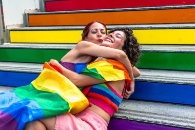 İki kadın sevgiyle kucaklaşıyor, gökkuşağı bayrağına sarılmış, canlı bir şekilde boyanmış merdivenlerde oturuyor, aşkı, gururu ve Igbtq artı hakları sembolize ediyor.