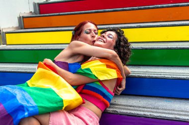 İki kadın kucaklaşıyor ve öpüşüyor, gökkuşağı bayrağına sarılmış, lgbtq renklerine boyanmış merdivenlerde artı gurur bayrağı, bir gurur etkinliği sırasında aşkı, çeşitliliği ve eşitliği kutluyorlar.