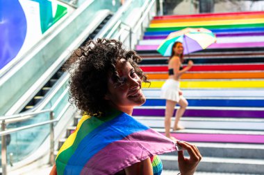 Gurur verici bir etkinlikte gökkuşağı bayrağı gösteren genç bir kadın, çeşitliliği kutlayan, lgbtq artı hakları ve renkli gökkuşağı merdivenleri ile kapsayacak bir kişi ve arka planda yürüyen başka bir kişi.