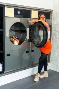 Genç bir kadın, çamaşırhanedeki açık bir çamaşır makinesine yaslanıyor. Çamaşırlarının yıkanmasını ve kurumasını bekliyor. Temiz ve yumuşak çamaşırların tadını çıkarıyor.