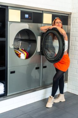 Çamaşırhanedeki açık bir kurutucuya yaslanan genç bir kadın, temiz giysiler içeride yuvarlanırken çamaşırları bekliyor, kolaylık ve verimliliği vurguluyor.