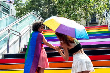 İki kadın, biri canlı gökkuşağı peleriniyle süslenmiş renkli gökkuşağı merdivenlerinde el ele tutuşmuş, lgbtq artı gururu kutluyor ve şehir ortamında çeşitliliği kucaklıyor.