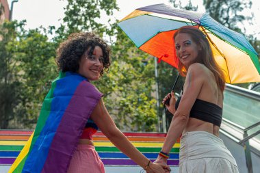 İki kadın el ele tutuşup yürüyorlar, gururu gökkuşağı bayrağı ve şemsiyeyle kutluyorlar, aşkı, çeşitliliği ve lgbtq artı hakları sembolize ediyorlar.