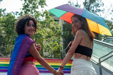 İki kadın el ele tutuşuyor ve gülümsüyor, biri gökkuşağı şemsiyesi tutuyor, gökkuşağı bayrağı ve makyajıyla gururu kutlarken, aşkı, çeşitliliği ve lgbtq artı hakları simgeliyor.