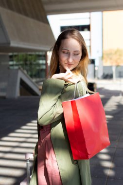 Şık bir turist şehir ortamında dolaşır, kırmızı alışveriş çantası taşır ve bavulu çeker, sonbaharda tatilde perakende terapinin keyfini çıkarır.