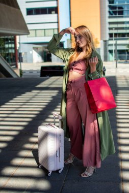 Tramvayı ve alışveriş çantalarıyla şehirde gezintiye çıkmayı beklerken gözlerini güneş ışığından koruyan genç bir kadın tatil boyunca güneşli bir günün tadını çıkarıyor.