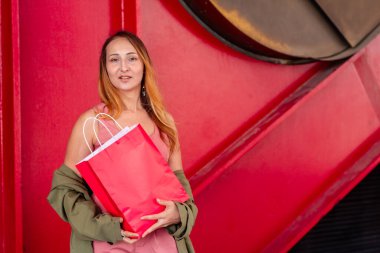 Büyük, canlı kırmızı bir alışveriş çantası tutan şık genç bir kadın çarpıcı bir kırmızı sanayi zeminine karşı poz verirken, tüketim ve kentsel tarzın bir karışımı olduğunu öne sürüyor.