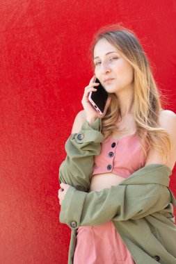 Akıllı telefon kullanan genç bir kadın, canlı kırmızı bir arkaplana karşı güvenle ayakta duruyor. Günlük kıyafetleri rahat ve modern bir yaşam tarzını çağrıştırıyor. Bağlılık ve stili vurguluyor.