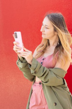 Pembe akıllı telefonuyla canlı kırmızı bir duvara yaslanmış zarif genç bir kadın, sonbahar sahnelerini yakalıyor ve sonraki macerasını çağdaş bir ortamda planlıyor.