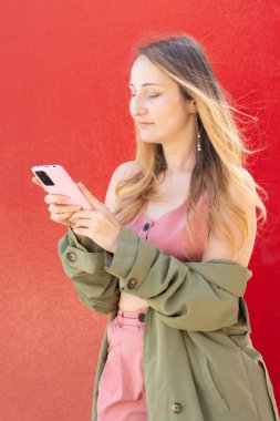 Pembe akıllı telefon kullanan genç bir kadın, açık pembe bir elbise ve yeşil bir ceket giyiyor, canlı kırmızı bir arka plana karşı duruyor, mobil teknolojinin tadını çıkarıyor.