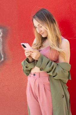 Pembe elbiseli ve yeşil ceketli şık genç bir kadın, sonbahar sezonunda canlı kırmızı bir duvara yaslanırken cep telefonunu kullanıyor.