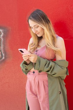 Havalı bir sonbahar gününde canlı kırmızı bir duvara yaslanırken akıllı telefonunu kullanan şık genç bir kadın, modaya uygun sonbahar kıyafetinin ve şehir yaşam tarzının altını çiziyor.