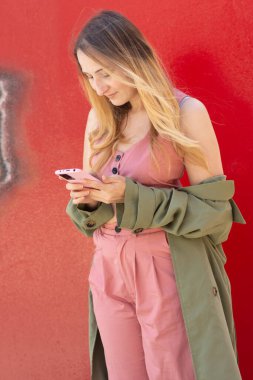 Şık bir sonbahar elbisesi içinde şık genç bir kadın, canlı kırmızı bir duvara karşı duruyor, akıllı telefonuna sarılmış, teknolojinin ve modern trendlerin kesişimlerini sergiliyor.